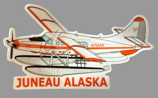 A die-cut sticker of a white and orange seaplane with the text 'JUNEAU ALASKA' below it, placed on a brown wooden surface.