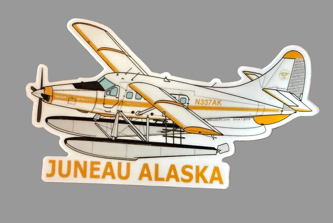 A die-cut sticker of a Juneau Alaska seaplane on a brown wood-grain background. The sticker features a white and yellow seaplane with the text 'JUNEAU ALASKA' in yellow at the bottom.