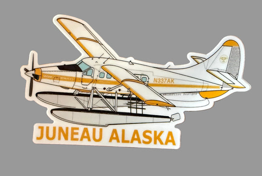 A die-cut sticker of a Juneau Alaska seaplane on a brown wood-grain background. The sticker features a white and yellow seaplane with the text 'JUNEAU ALASKA' in yellow at the bottom.